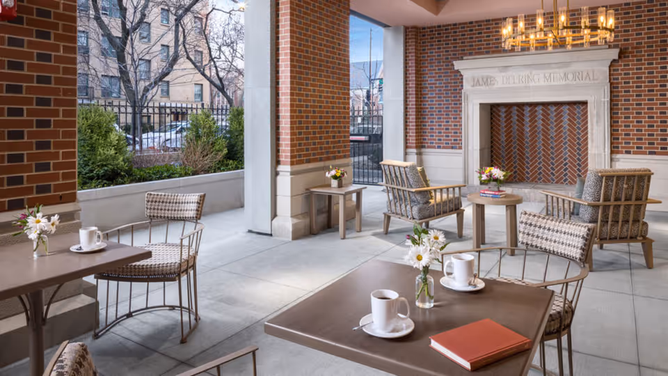 Outdoor covered patio area with brick walls and a decorative fireplace labeled 'James Delring Memorial'. The space features several tables and chairs with cushions, small vases of flowers, coffee cups, and a book on one table. There are shrubs and trees visible outside the fenced area.