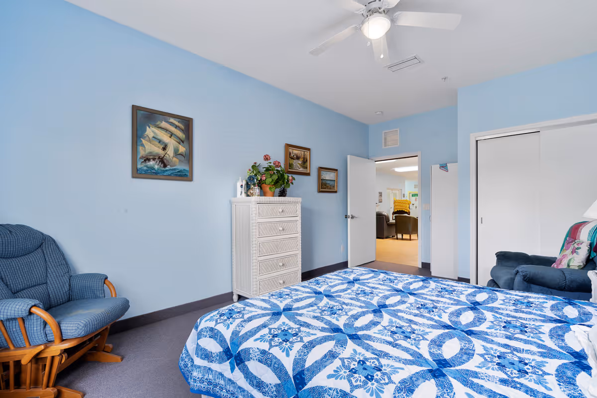 A bedroom with light blue walls featuring a bed with a blue and white patterned quilt, a blue cushioned rocking chair, a white chest of drawers with a potted plant and decorative items on top, and framed paintings on the wall. The door is open, showing a glimpse of a living area with chairs and a yellow pillow.