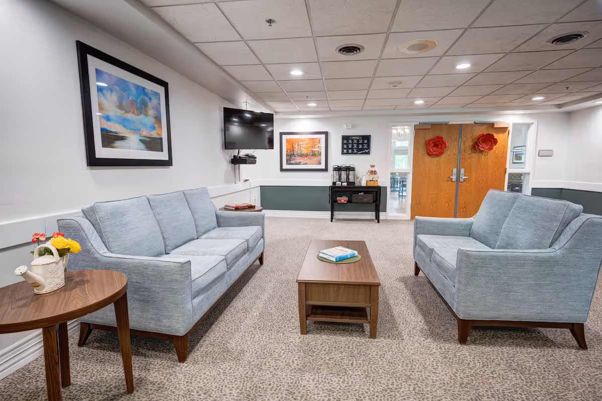 A cozy common area with two light blue sofas facing each other across a wooden coffee table with a book on it. A small round side table with a white watering can and yellow flowers is beside one sofa. The room has carpeted flooring with a subtle pattern, white walls with a green lower panel, and a ceiling with recessed lighting. On the walls are two framed paintings and a mounted flat-screen TV. In the background, double wooden doors decorated with red flower ornaments lead to another room.