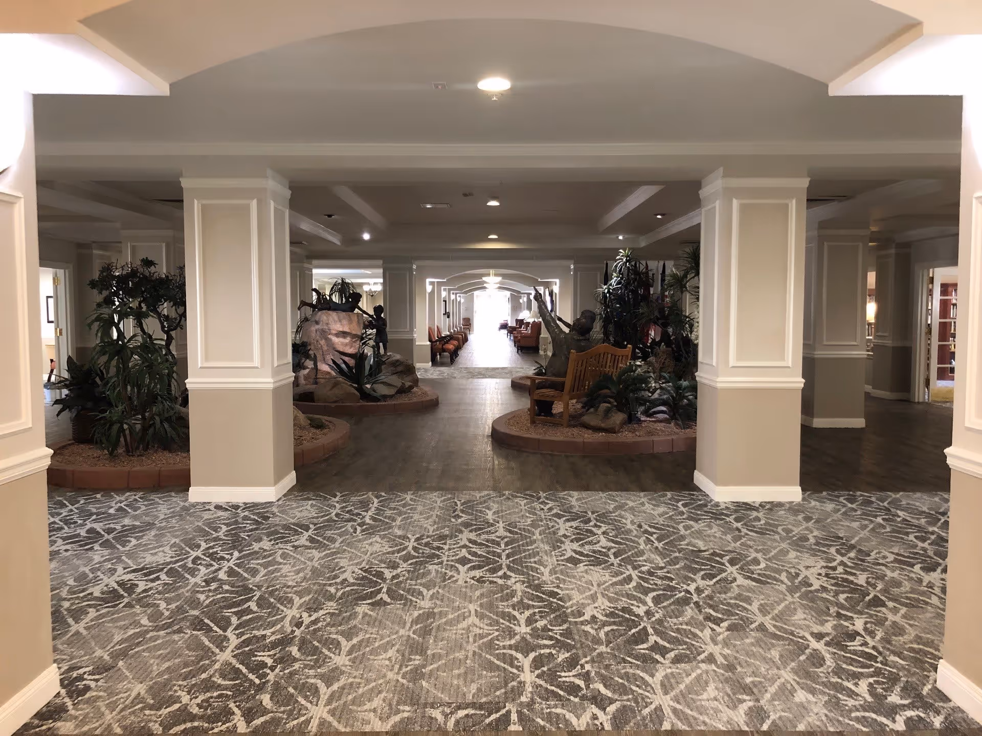 Interior view of a senior living facility hallway with beige walls, white pillars, patterned carpet, and wooden flooring. There are indoor plants and a wooden bench with a sculpture of a person sitting on it. The hallway extends into a seating area with chairs and tables in the distance.