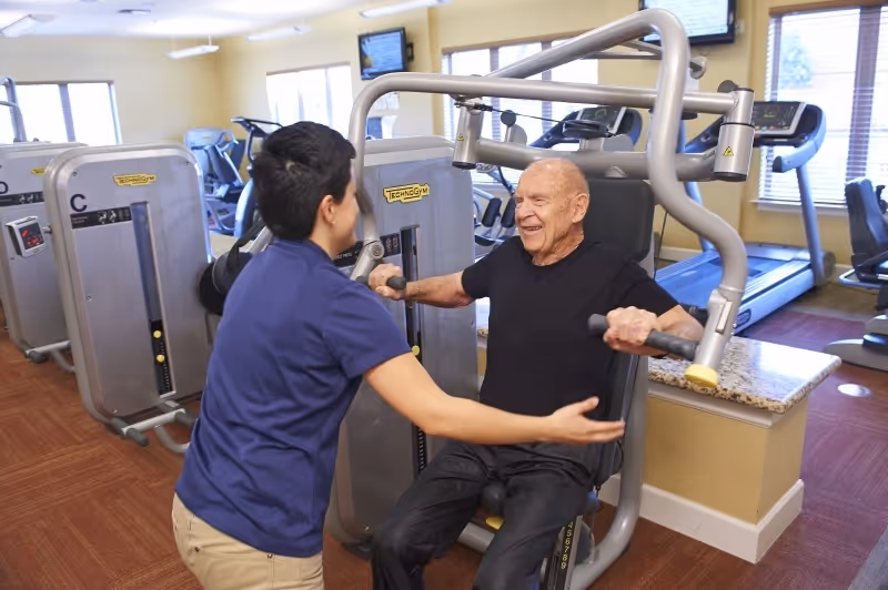 An elderly man using a chest press exercise machine in a fitness room, assisted by a staff member wearing a blue shirt. The room has exercise equipment including treadmills and weight machines, with windows letting in natural light.
