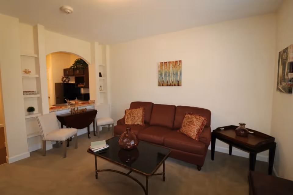 A cozy living room with a brown leather sofa adorned with two patterned cushions, a glass coffee table with a decorative vase and books, two white upholstered chairs near a small wooden table, and an open archway leading to a kitchen area with dark cabinets and a refrigerator. The walls are light-colored with two pieces of artwork hanging.