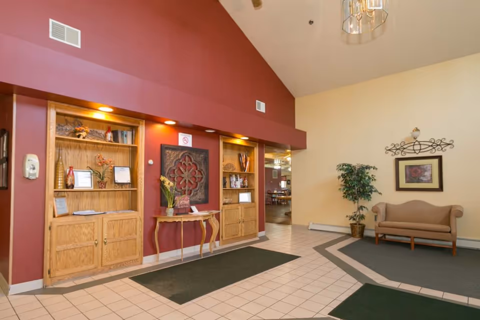 Interior view of a senior living facility lobby with beige and maroon walls, built-in wooden shelves with decorative items, a small table with a flower vase, a beige couch, a potted plant, framed artwork, and tiled floor with green mats.
