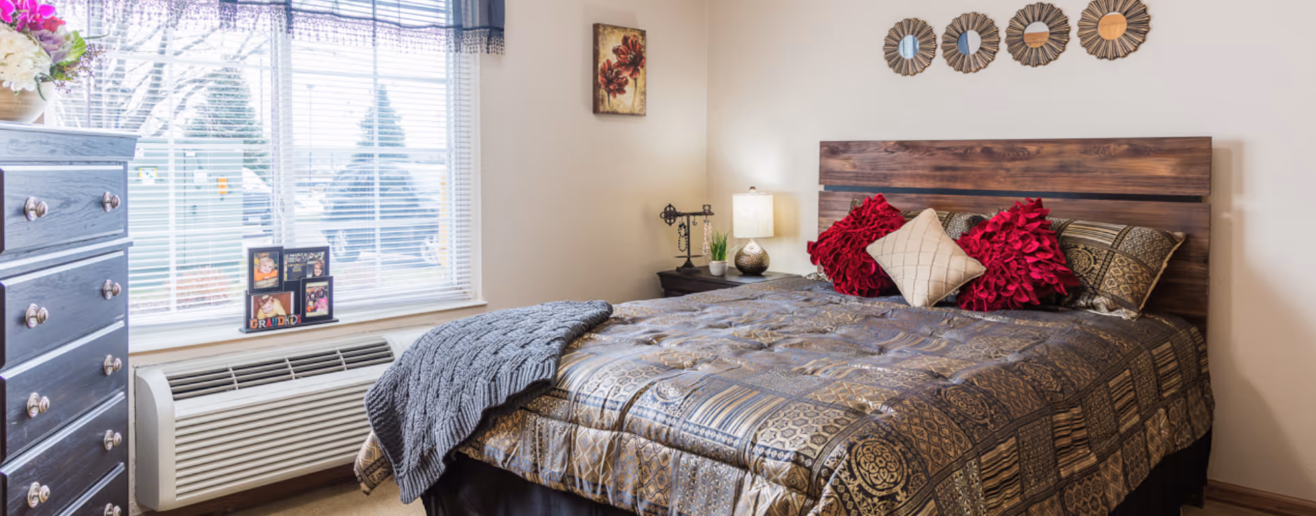 A cozy bedroom with a wooden headboard bed covered in a patterned comforter and decorative pillows in red and beige. A gray knitted throw blanket is draped over the bed. Next to the bed is a nightstand with a lamp, a small plant, and a decorative item. A window with blinds and a valance lets in natural light, and a dresser with a vase of flowers and framed photos is visible on the left side of the room. The walls are light-colored with a small piece of artwork and decorative mirrors above the bed.