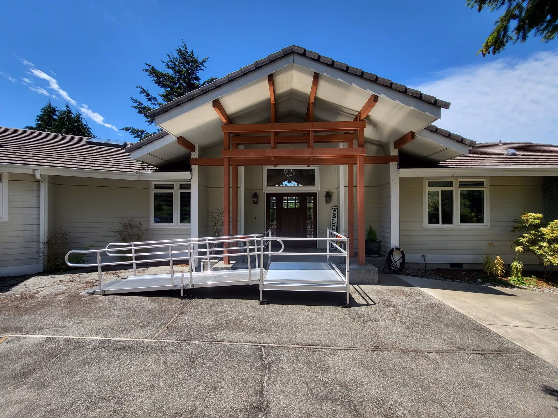 Front entrance of a single-story building with a peaked roof and wooden beams. There is a metal wheelchair ramp leading up to the door. The building is light-colored with several windows and some small plants near the entrance. The sky is clear with a few clouds.