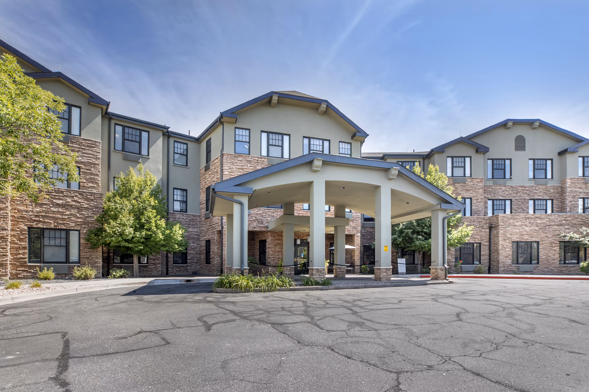 Exterior view of a three-story senior living facility building with a covered entrance, stone and beige siding, multiple windows, and some trees around the entrance area under a clear blue sky.