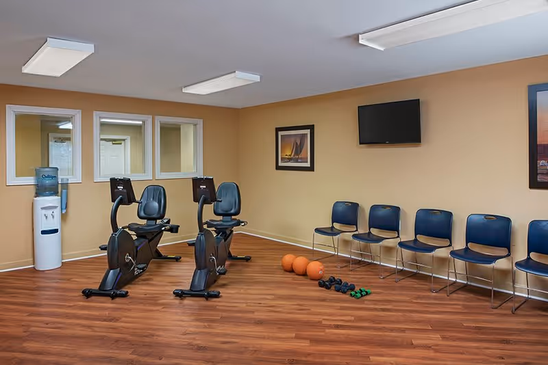 A small exercise room with two stationary recumbent bikes, a water cooler, several blue chairs lined up against a beige wall, three orange exercise balls, and a set of dumbbells on a wood floor. The walls have framed pictures and windows looking into another room.