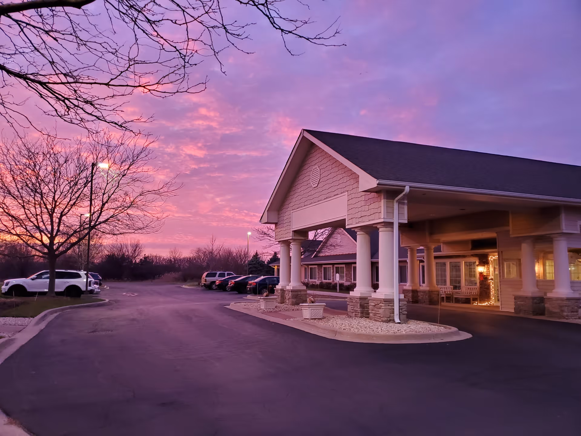 Exterior view of Rock Run Memory Care facility at dusk with a purple and pink sky. The building has a covered entrance supported by white columns and stone accents. Several parked cars and leafless trees are visible in the parking lot.