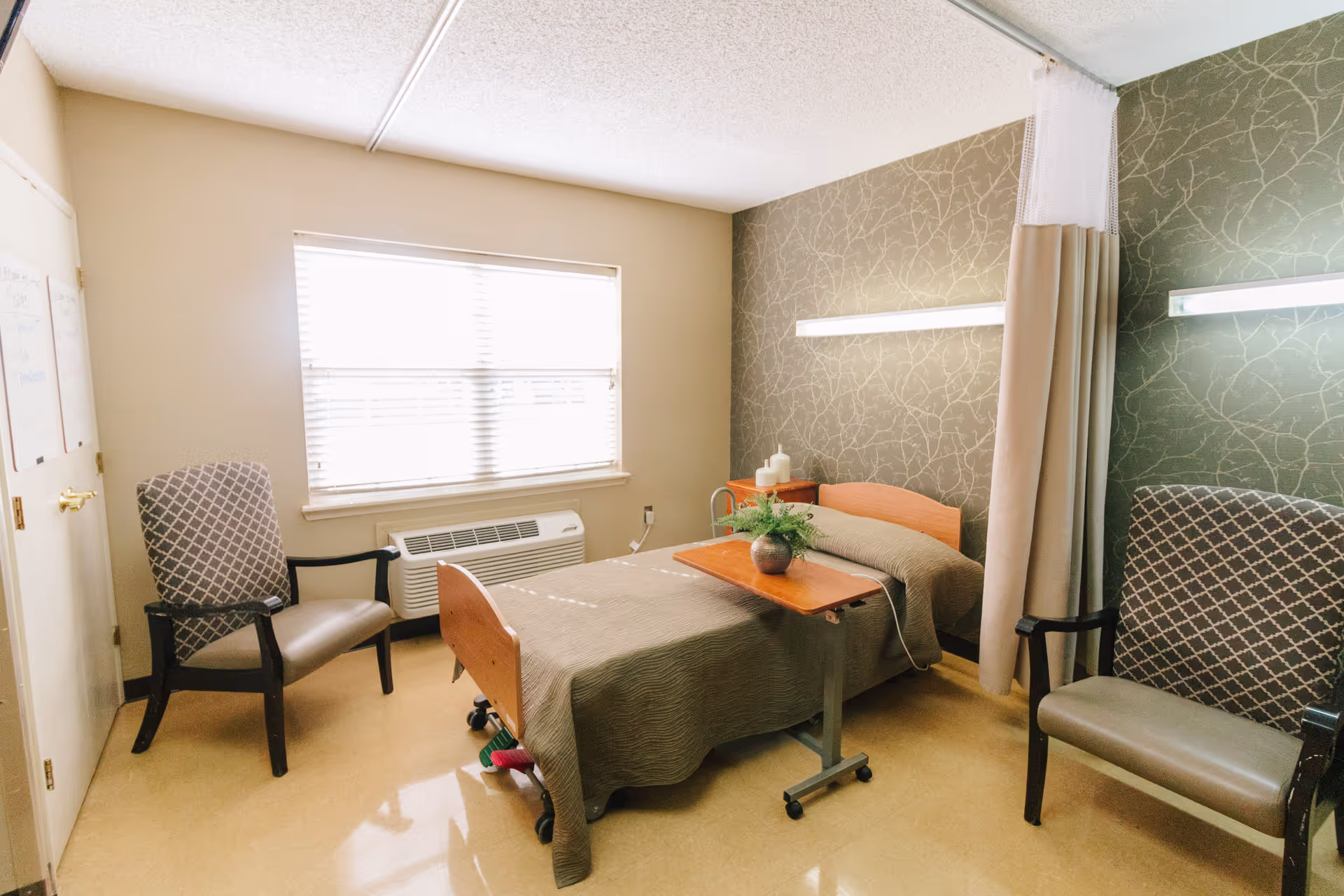 A small, well-lit room with a single hospital-style bed covered with a brown quilt. There is a wooden overbed table with a small plant on it. Two cushioned chairs with patterned upholstery are placed on either side of the bed. A window with blinds lets in natural light, and a beige curtain hangs partially around the bed. The walls are painted beige with one accent wall featuring a subtle branch pattern. There is an air conditioning unit below the window.