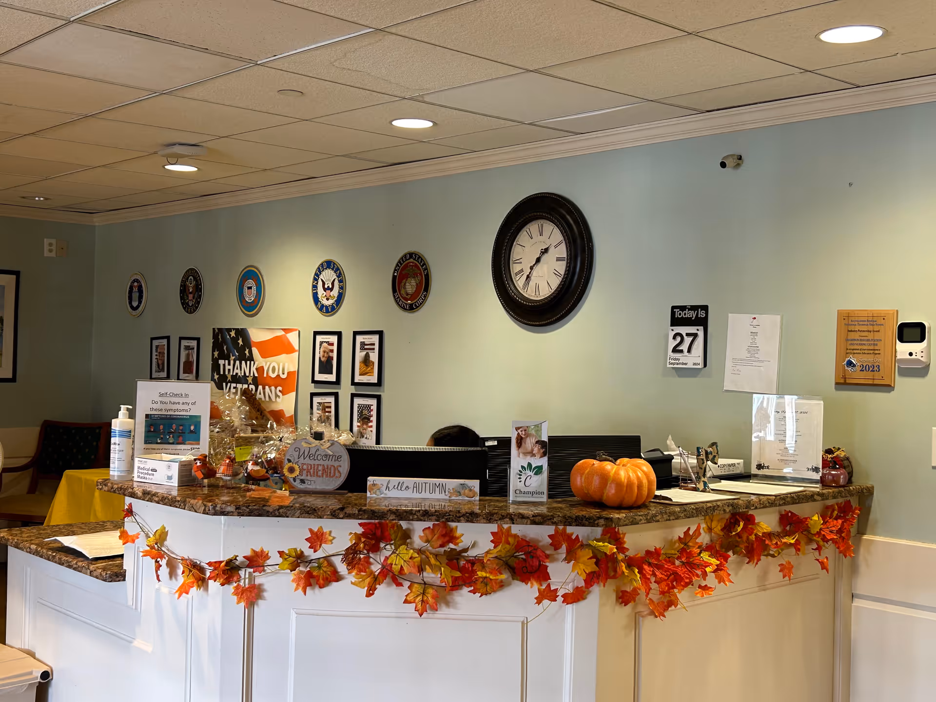 Reception desk in a nursing facility decorated for autumn with garlands, a pumpkin, a wall clock and veterans' plaques.