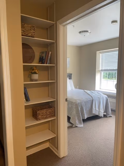 View of a bedroom through an open doorway, showing a neatly made bed with a light-colored bedspread and a window with blinds letting in natural light. To the left of the doorway, there is a built-in shelving unit with decorative items including books, a basket, a small plant, and a round tray.