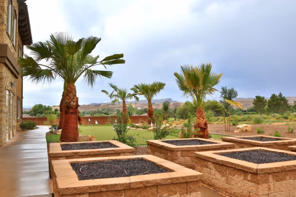 Outdoor area at The Retreat at Sunbrook featuring a paved walkway alongside a building, several palm trees, raised garden beds with soil, and a putting green with flags in the background under a cloudy sky.