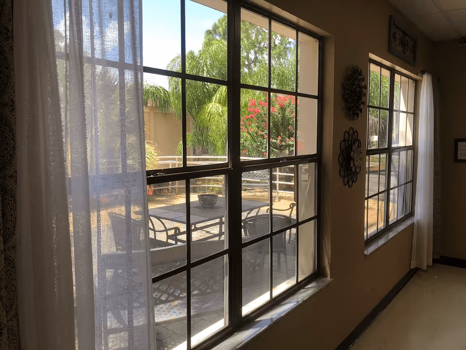 View through large windows with black frames showing an outdoor patio area with a table and chairs, surrounded by greenery including palm trees and flowering bushes. Sheer white curtains hang on either side of the windows inside a room with beige walls decorated with metal wall art.