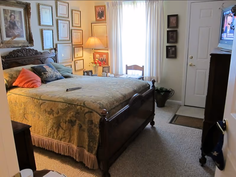 Cozy furnished bedroom with a large wooden bed, framed pictures on the wall, a nightstand lamp, and a window with curtains.