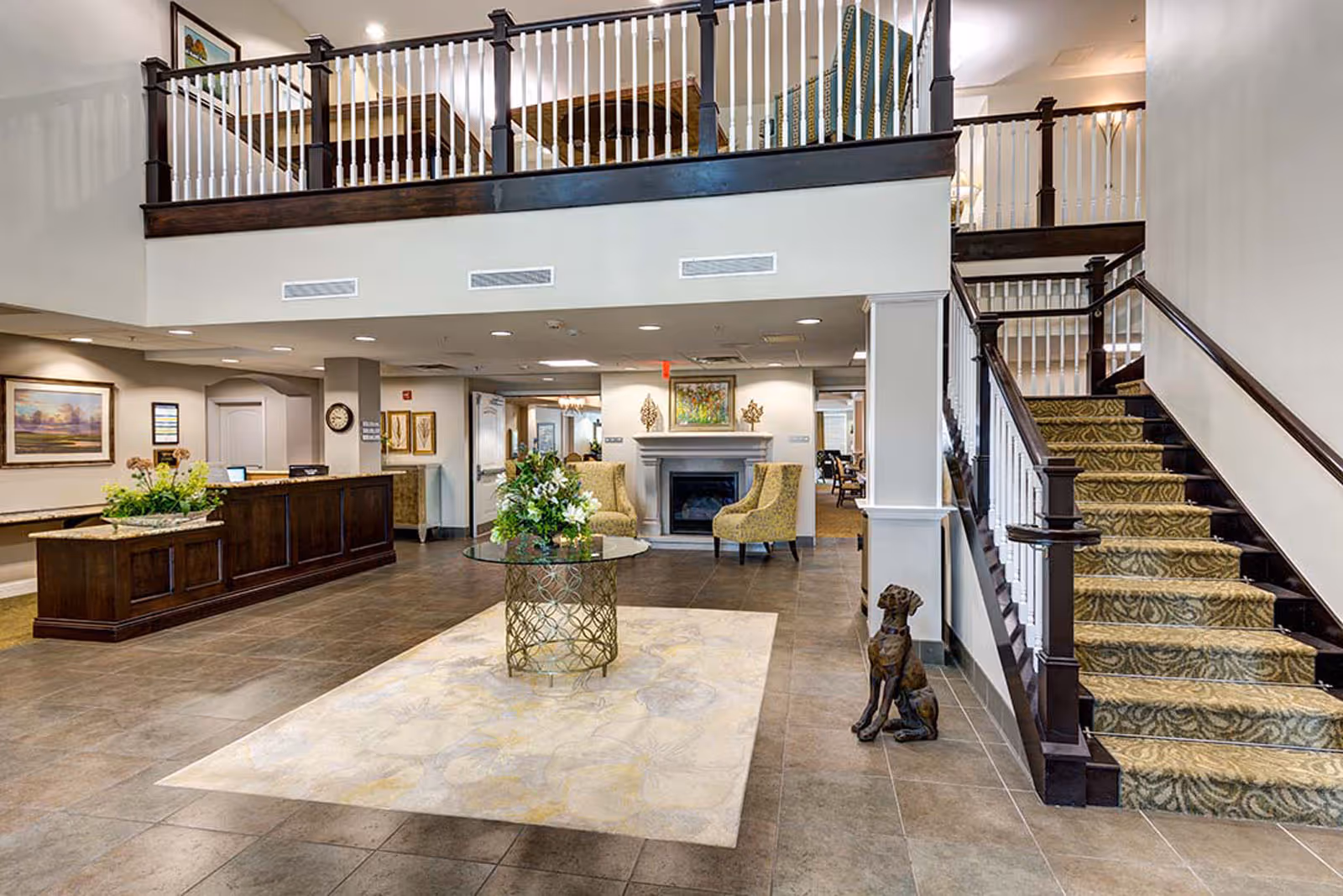 Spacious and well-lit senior living facility lobby with a tiled floor, a central glass table with a floral arrangement, a fireplace flanked by two patterned armchairs, a wooden reception desk on the left, and a carpeted staircase with dark wood railing on the right. There is a decorative dog statue at the base of the stairs and a balcony with white spindles overlooking the lobby.