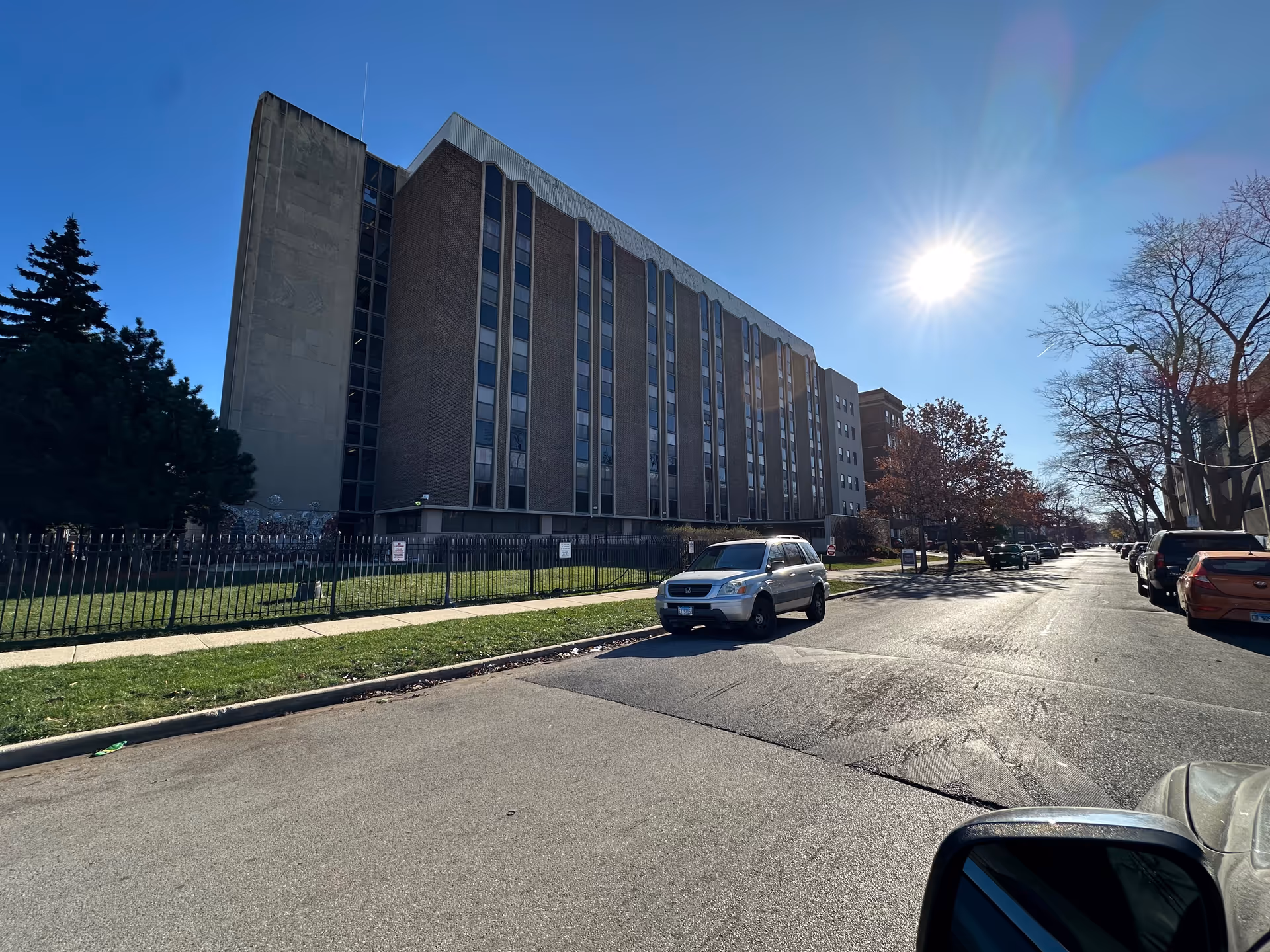Large multi-story brick apartment building on a sunny street with parked cars and a fenced grassy front yard.