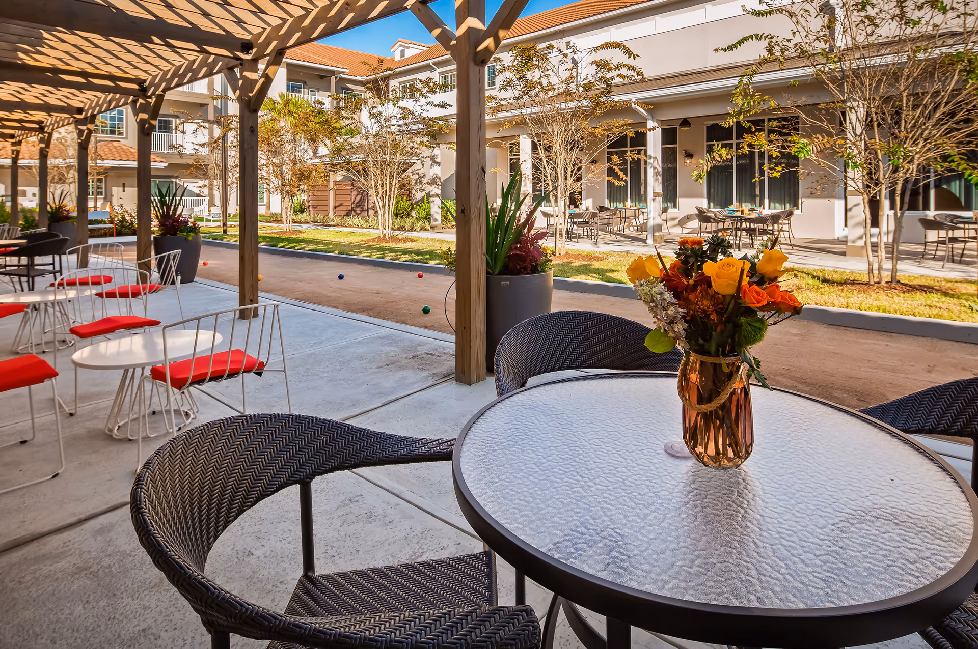 Outdoor covered courtyard seating area with tables and chairs, a vase of flowers on a table, and a bocce court in front of a senior living building.