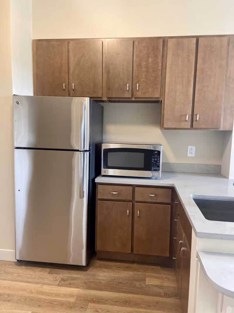 A kitchen corner featuring a stainless steel refrigerator, a microwave oven on a countertop, wooden cabinets above and below the counter, and a sink on the right side. The floor is wooden and the walls are light-colored.
