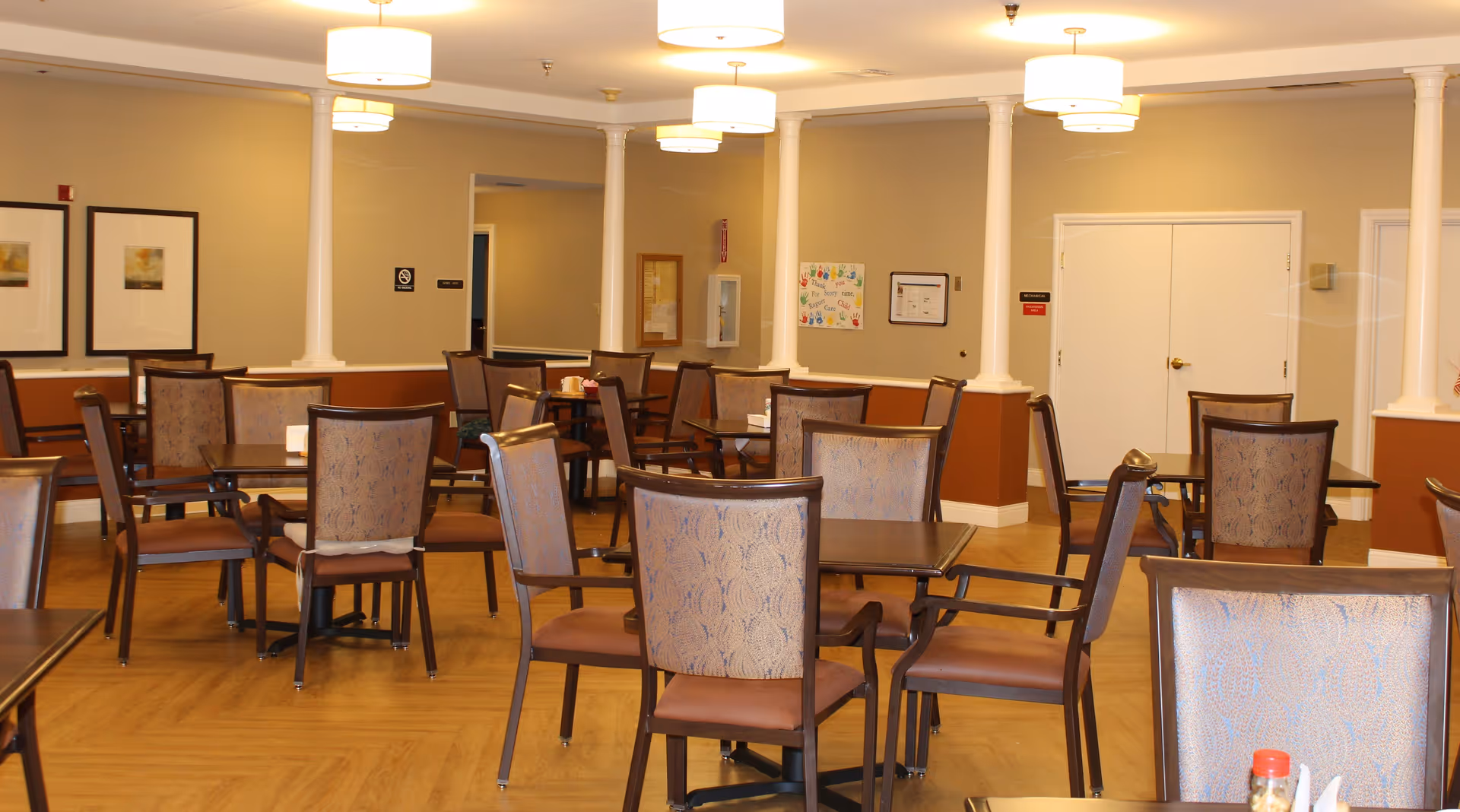 Interior view of a dining room in an assisted living facility with multiple tables and chairs arranged neatly. The room has beige walls with white columns, framed artwork on the walls, and ceiling lights providing warm illumination.
