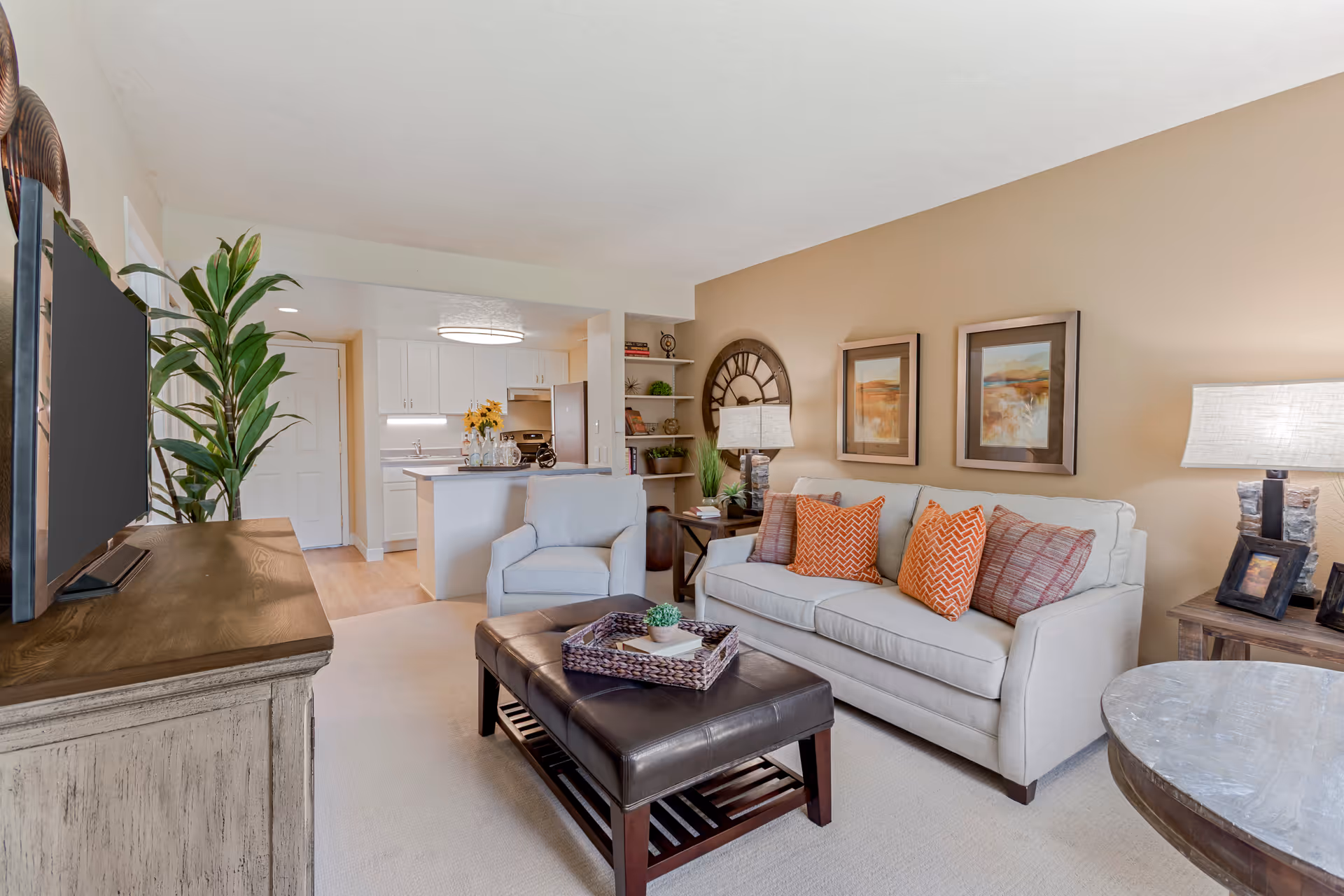 A cozy living room with a beige sofa adorned with orange and patterned pillows, a matching armchair, and a dark brown leather ottoman coffee table. The room features a wooden TV stand with a flat-screen TV, a tall green plant, and a side table with a lamp and picture frames. In the background, a kitchen area with white cabinets, a refrigerator, and a countertop with a vase of sunflowers is visible. The walls are painted a warm beige color and decorated with framed artwork and a large clock.