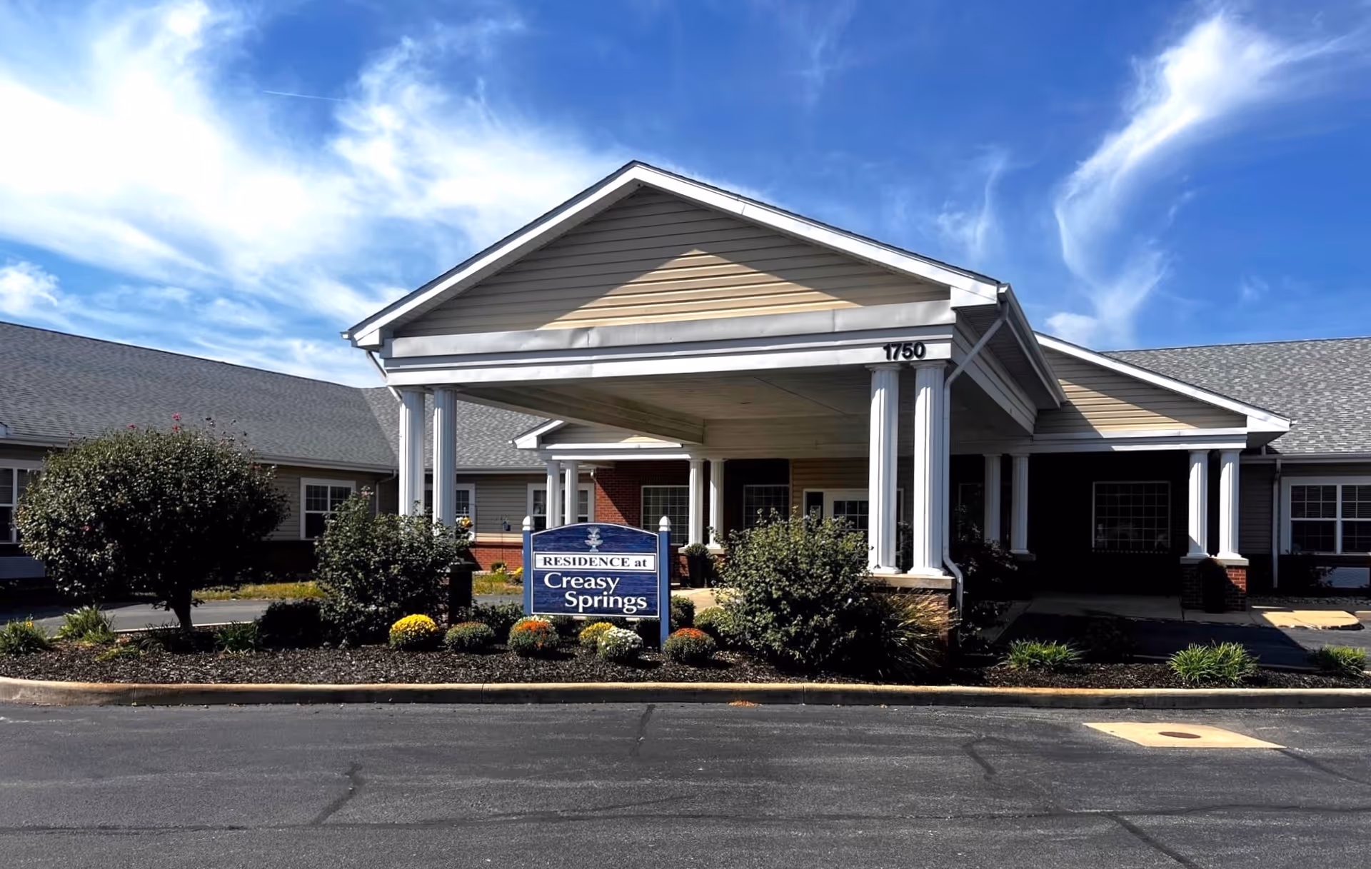 Front exterior view of Creasy Springs Health Campus building with a covered entrance supported by white columns, landscaped bushes and flowers in front, and a blue sign that reads 'Residence at Creasy Springs'. The sky is partly cloudy with blue and white clouds.