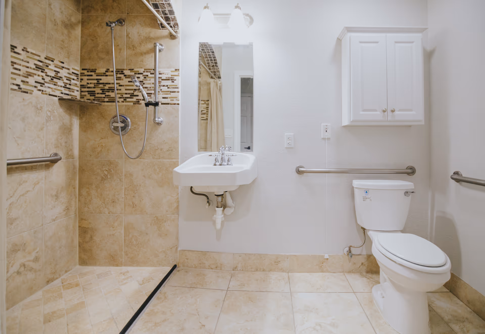 A clean, accessible bathroom with beige tiled floors and walls. The bathroom features a walk-in shower with a handheld showerhead, a white wall-mounted sink with a mirror above it, a white toilet, and white storage cabinet mounted on the wall. There are stainless steel grab bars next to the toilet and in the shower area for support.