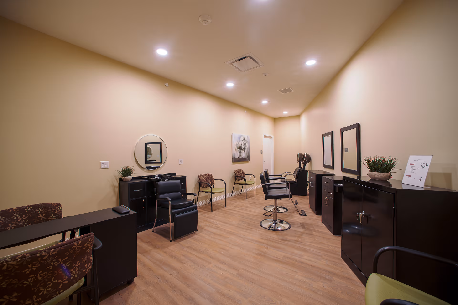Interior view of a senior living facility's salon room with beige walls and wooden flooring. The room contains black salon chairs, mirrors mounted on the walls, small black cabinets, and a few chairs with patterned upholstery. There are potted plants on the cabinets and a framed artwork on the wall.