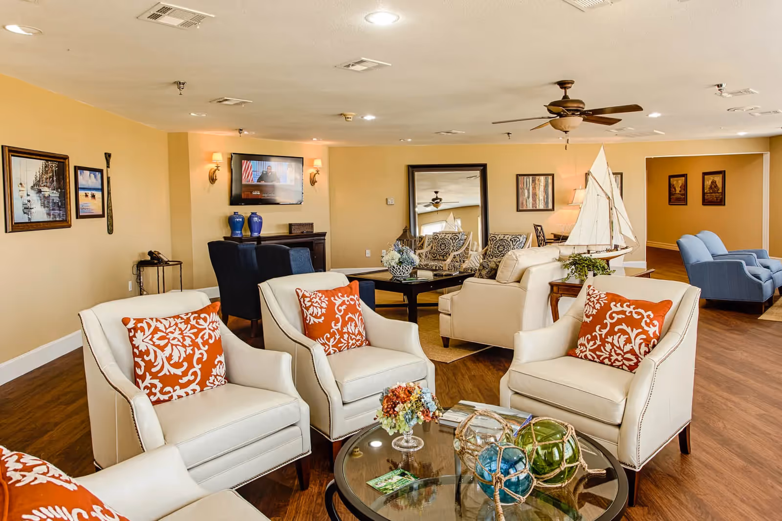 A cozy senior living facility lounge with cream-colored armchairs featuring orange patterned cushions arranged around a glass coffee table decorated with colorful glass floats and a small floral arrangement. The room has warm yellow walls adorned with framed paintings, a ceiling fan, a large mirror, and a wall-mounted TV. Additional seating includes blue armchairs and a beige sofa, with decorative items such as a model sailboat and blue vases on a sideboard.