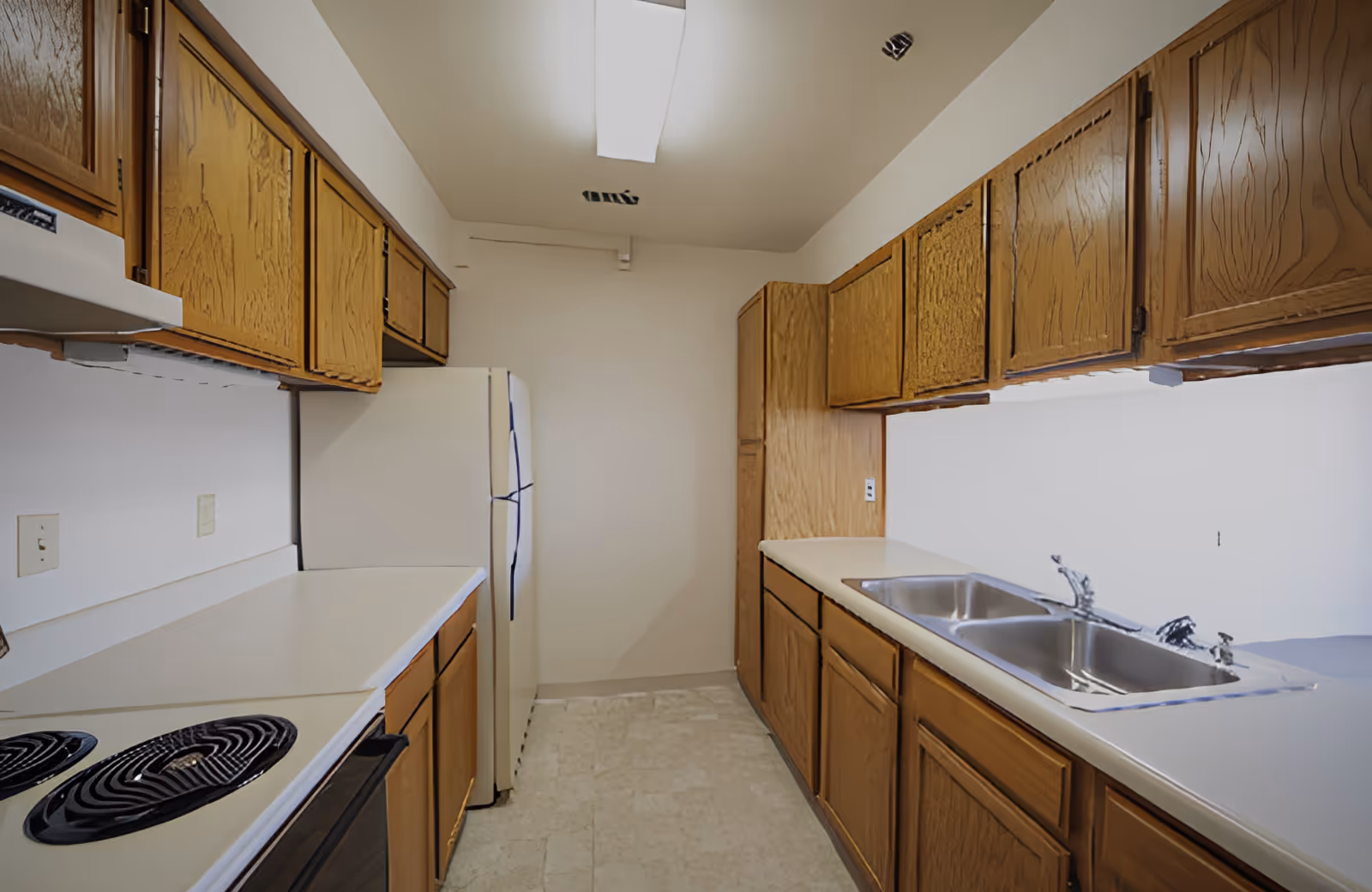 A narrow kitchen with wooden cabinets on both sides, a white refrigerator at the end, a double stainless steel sink on the right countertop, and an electric stove on the left countertop. The kitchen has a white ceiling with a fluorescent light fixture.