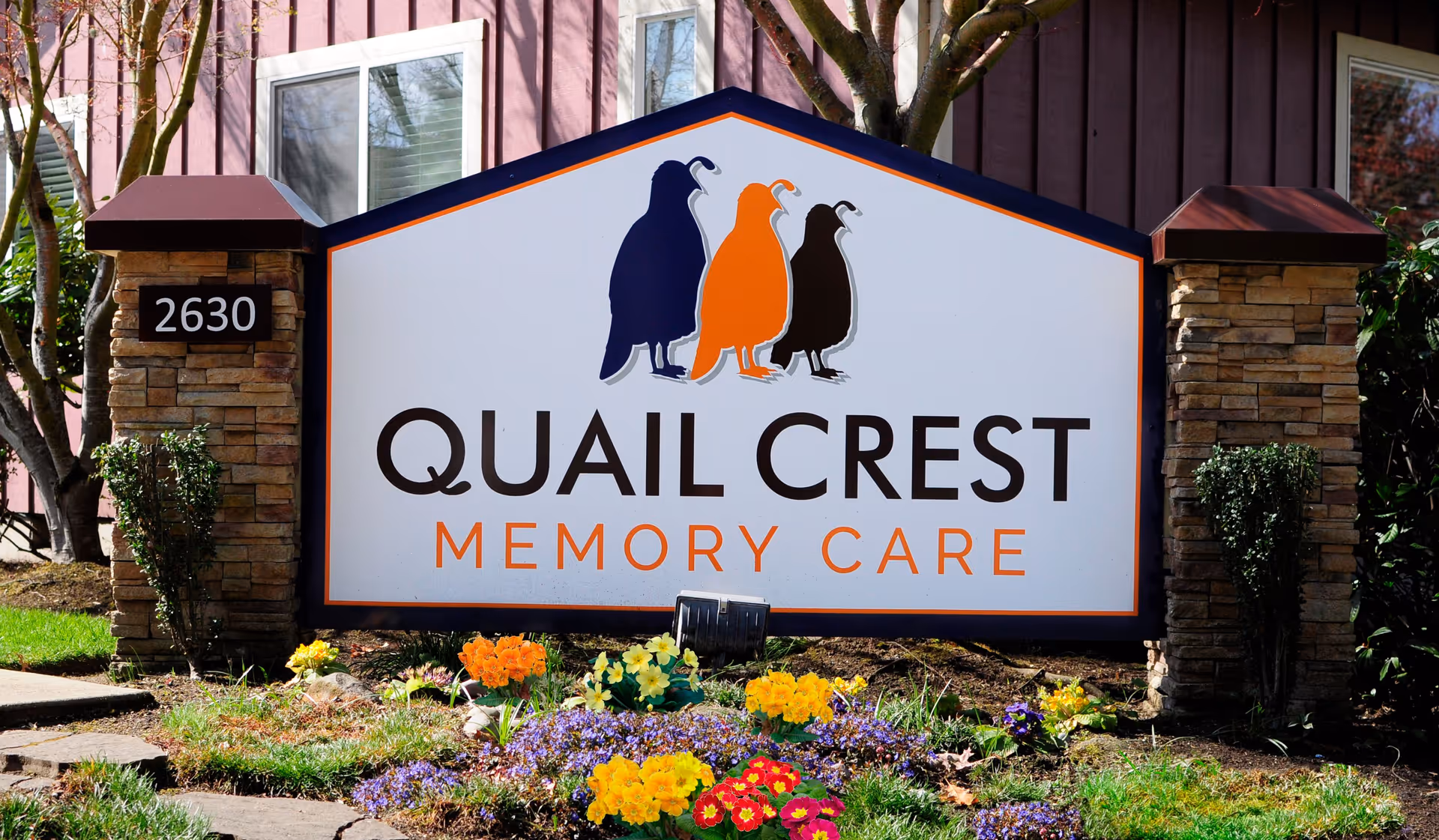 Outdoor entrance sign reading 'Quail Crest Memory Care' with three quail silhouettes, the address '2630', and flowers in front of a building.