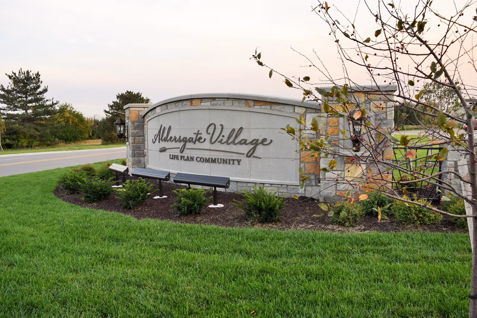 Stone entrance sign for Aldersgate Village Life Plan Community surrounded by green grass, small bushes, and a tree with some leaves. A road and trees are visible in the background under a cloudy sky.