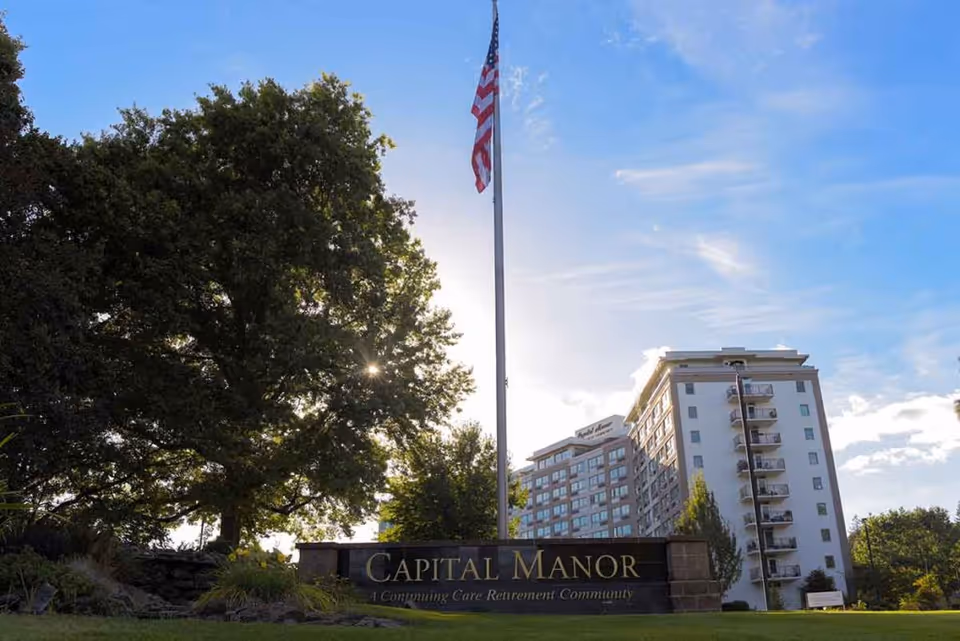 Exterior view of Capital Manor, a continuing care retirement community, showing a multi-story building with balconies, a large tree, an American flag on a flagpole, and a sign with the facility name in front.