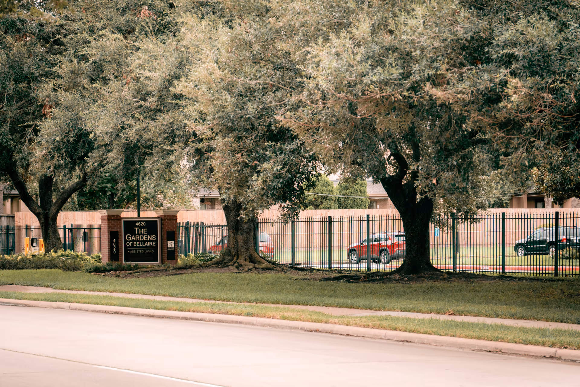 View of the entrance sign for The Gardens of Bellaire assisted living facility, surrounded by trees and a black metal fence, with parked cars visible in the background.