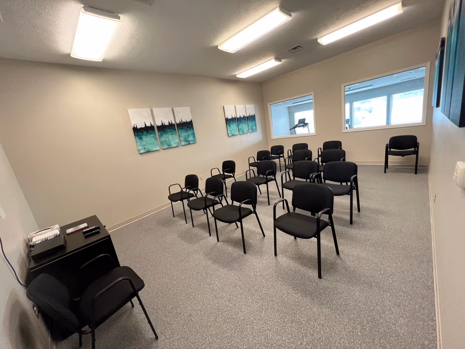 A small conference or meeting room with multiple black chairs arranged in rows facing a wall with three-panel abstract artwork. The room has gray carpet, white walls, and two windows looking into another room with exercise equipment. There is a small black table with some items on it in the corner.