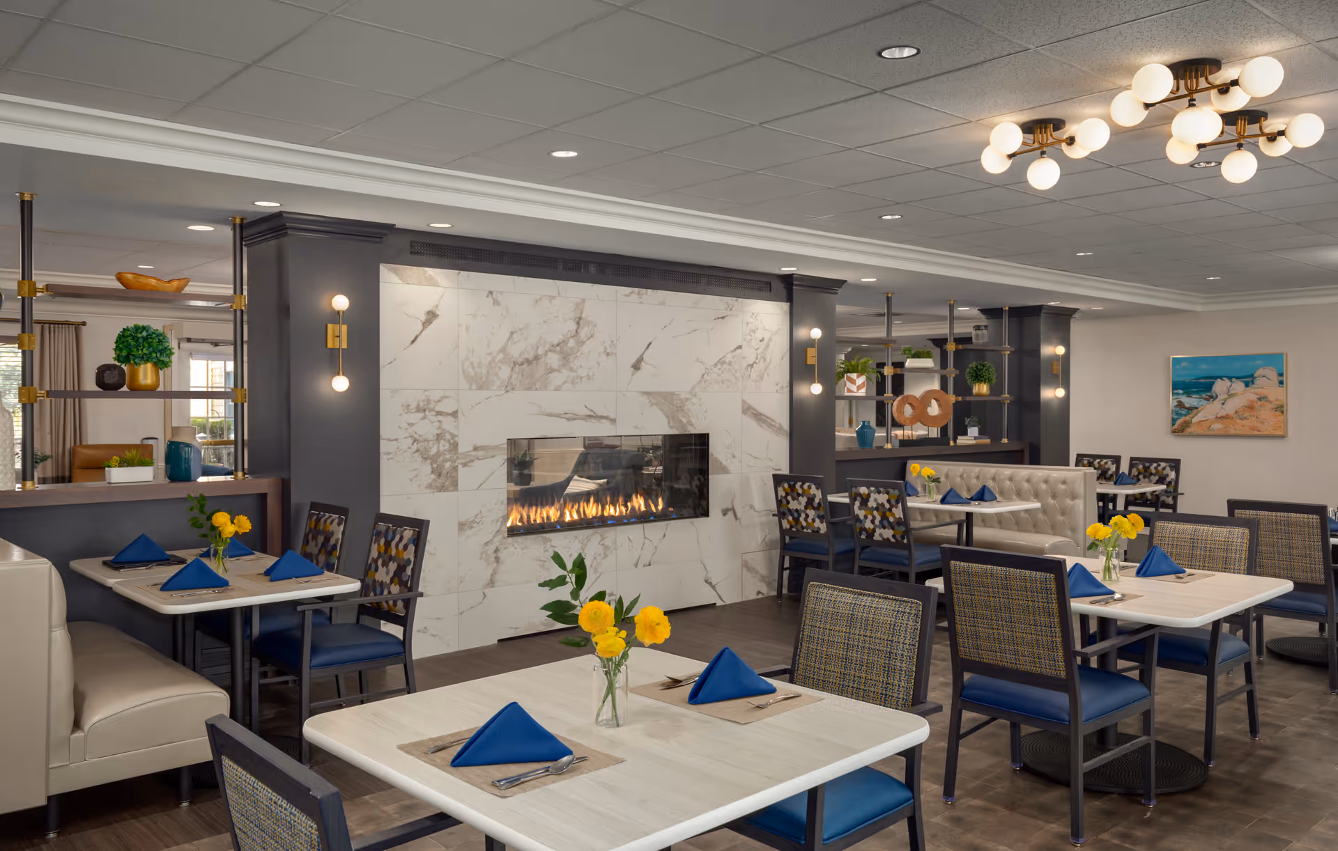 A modern dining room with several tables set with blue napkins and yellow flower centerpieces. The room features a large marble fireplace in the center with a gas fire, surrounded by dark columns and decorative shelving with plants and ornaments. The ceiling has recessed lighting and two modern chandeliers with globe bulbs. There is a painting of a coastal scene on the far wall.