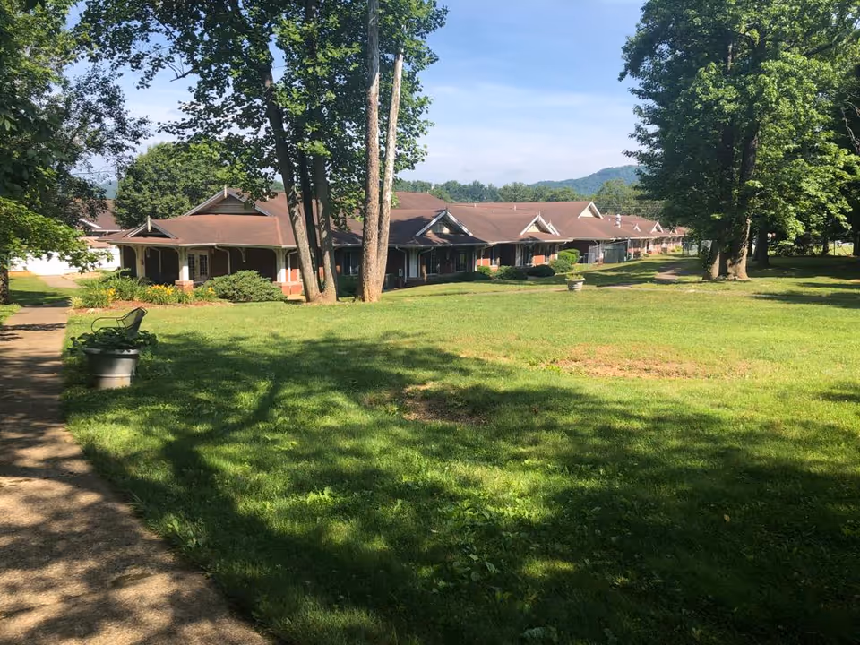 A large green lawn with several tall trees casting shadows, a paved walkway on the left side, and a long single-story building with a brown roof and covered porch in the background under a clear blue sky.