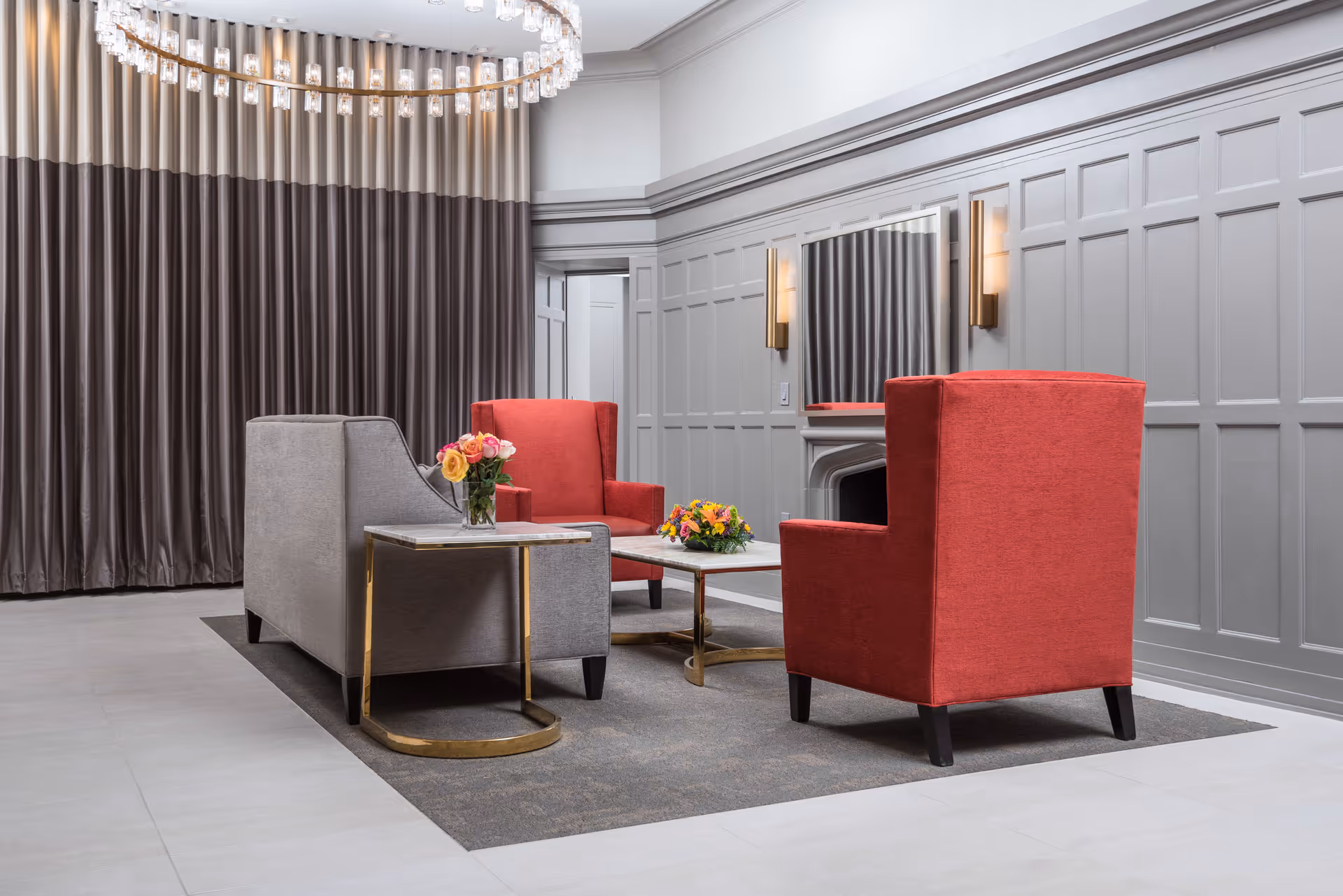 A modern and elegant sitting area with two red armchairs and one gray sofa arranged around a rectangular coffee table with floral arrangements. The room features gray paneled walls, a large mirror above a fireplace, wall sconces, and a large circular chandelier hanging from the ceiling. Floor-to-ceiling curtains in shades of gray cover the back wall.