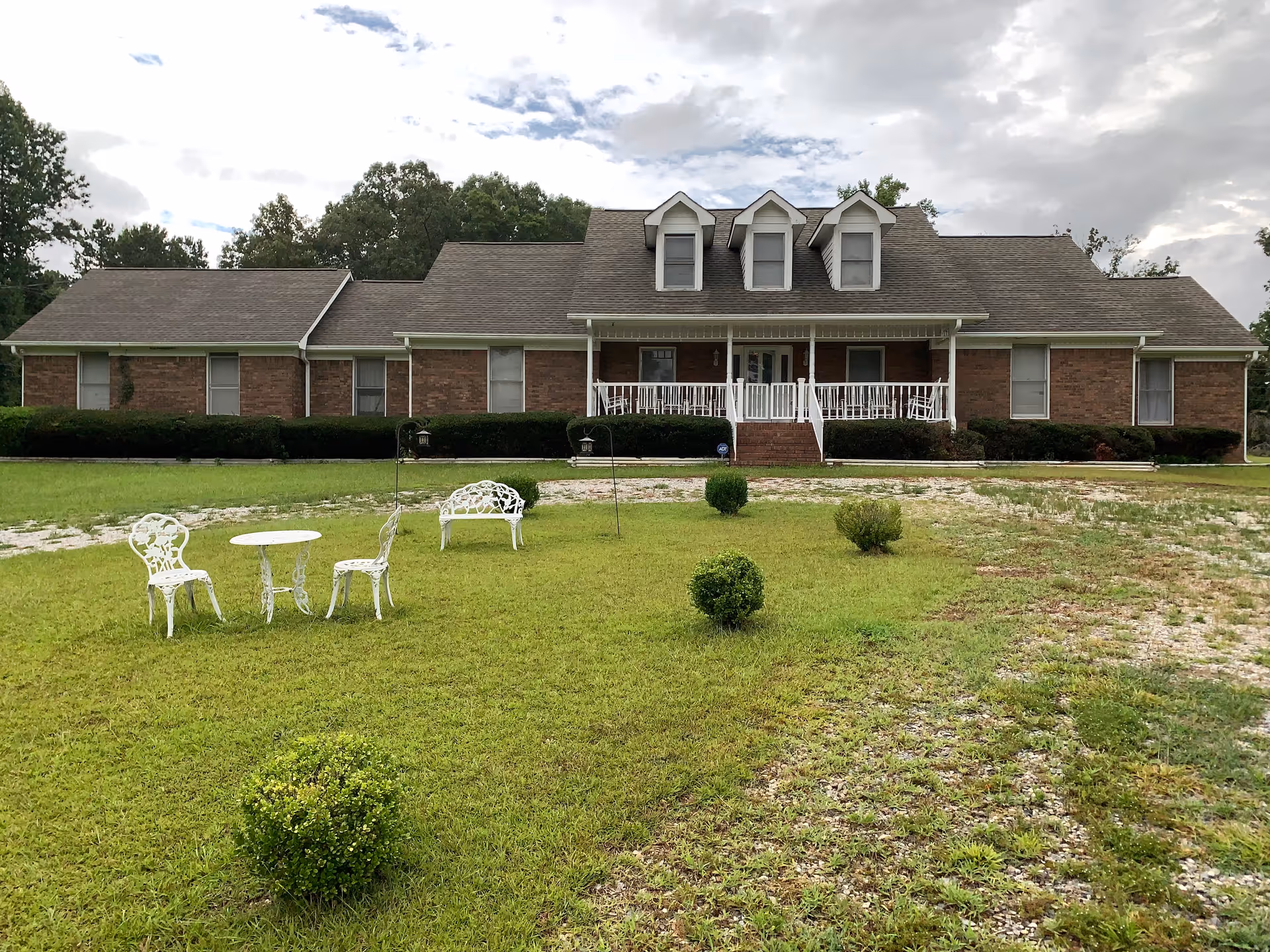 A large single-story brick building with a gray shingled roof and three dormer windows. The building has a white porch with railings and steps leading down to a grassy yard. In the yard, there are white metal outdoor chairs, a table, and a bench. Small bushes are scattered around the lawn, and trees are visible in the background under a cloudy sky.
