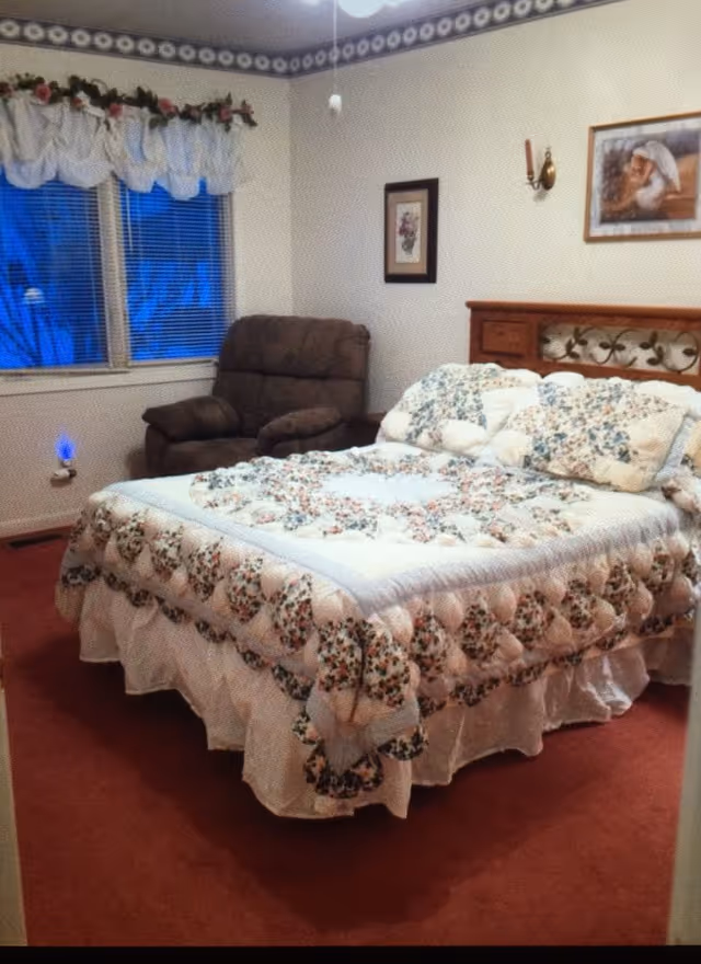 A bedroom with a bed covered in a floral quilt and matching pillows. There is a brown recliner chair next to a window with white curtains and floral valances. The walls are decorated with a floral border near the ceiling, a framed picture, and a wall-mounted candle holder.