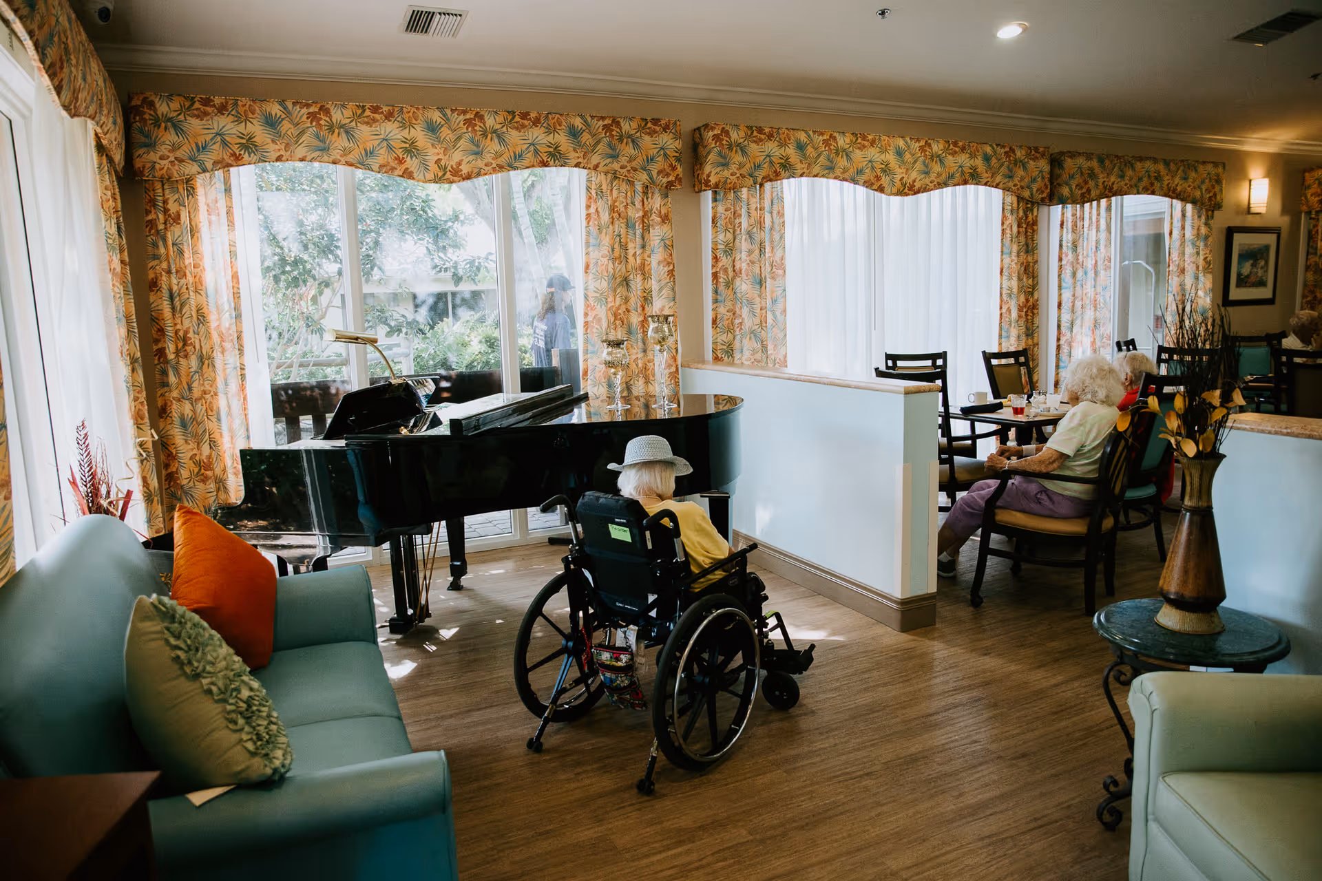 Senior residents relax in a bright common living area with a piano, a wheelchair, and seating.