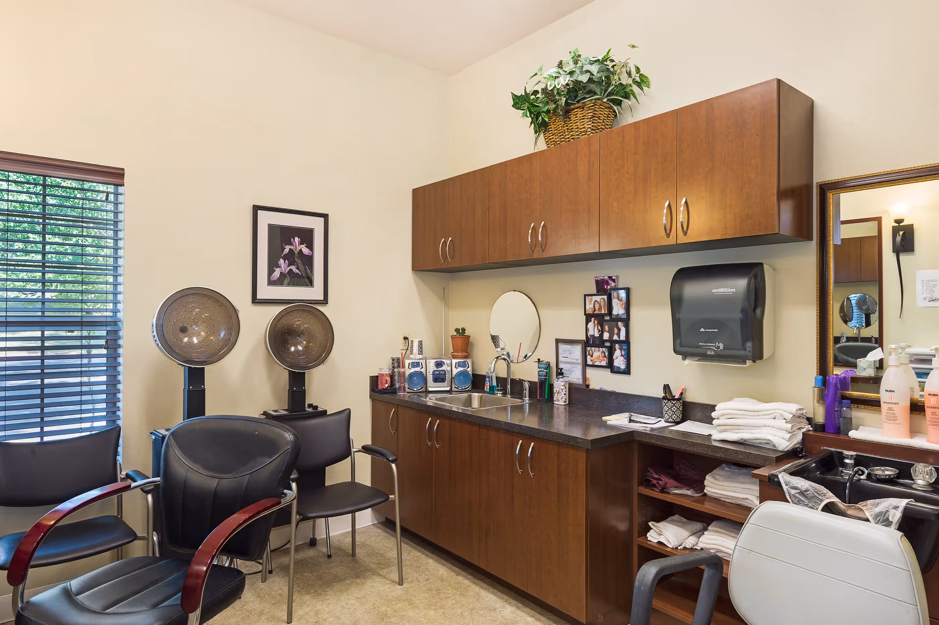 A well-lit salon/styling room with salon chairs, hooded hairdryers, a sink, wooden cabinets, and neatly stacked towels.