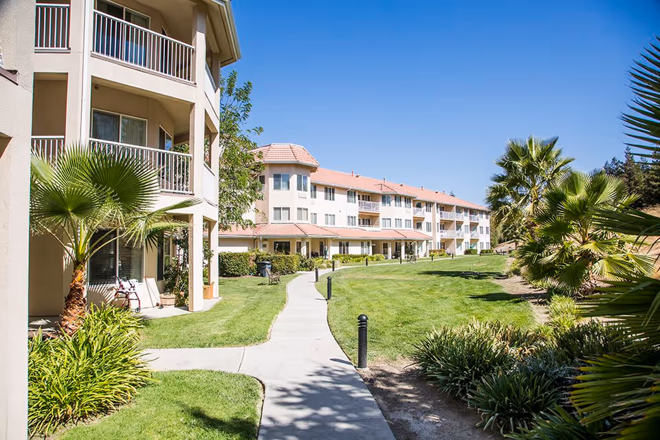 Outdoor view of a senior living facility with a paved walkway surrounded by green grass, palm trees, and shrubs. The building has multiple floors with balconies and a red-tiled roof under a clear blue sky.