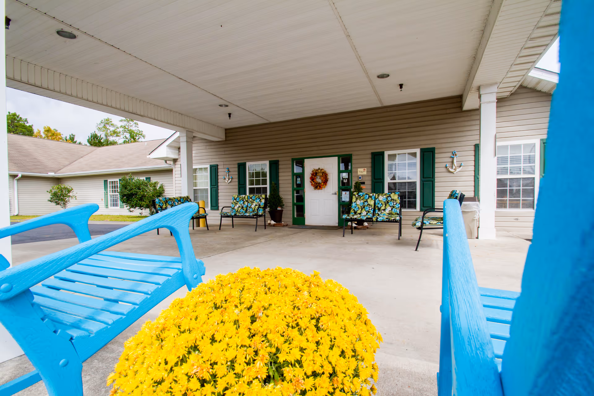 Covered entrance area of a building with beige siding and green shutters. There are blue wooden benches and floral cushioned chairs arranged on the concrete porch. A large pot of bright yellow flowers is in the foreground. The entrance door has a decorative wreath, and there are anchor decorations on the walls beside the windows.