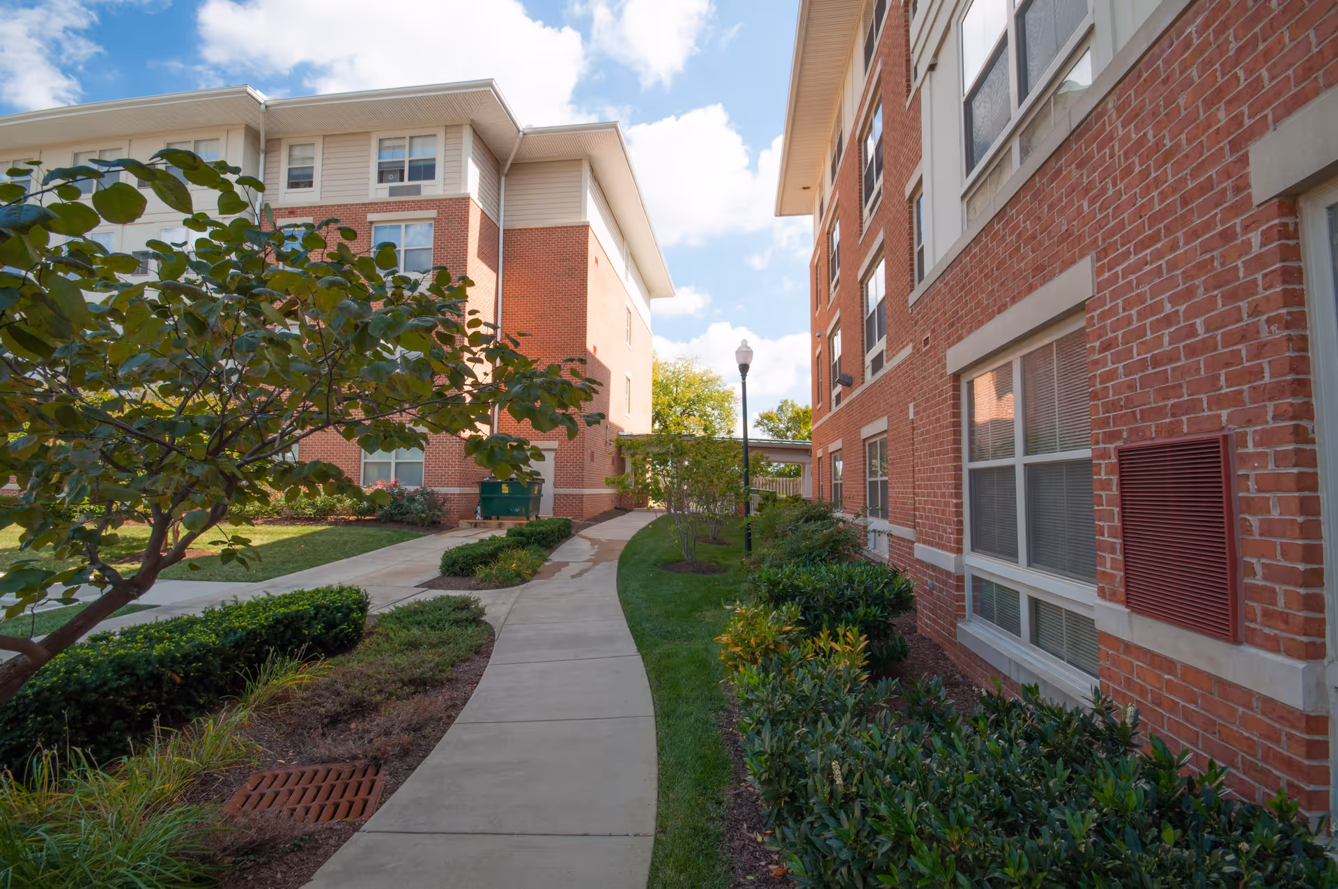 A paved walkway between two red brick apartment buildings with white trim. The path is surrounded by green bushes, small trees, and grass under a partly cloudy sky.