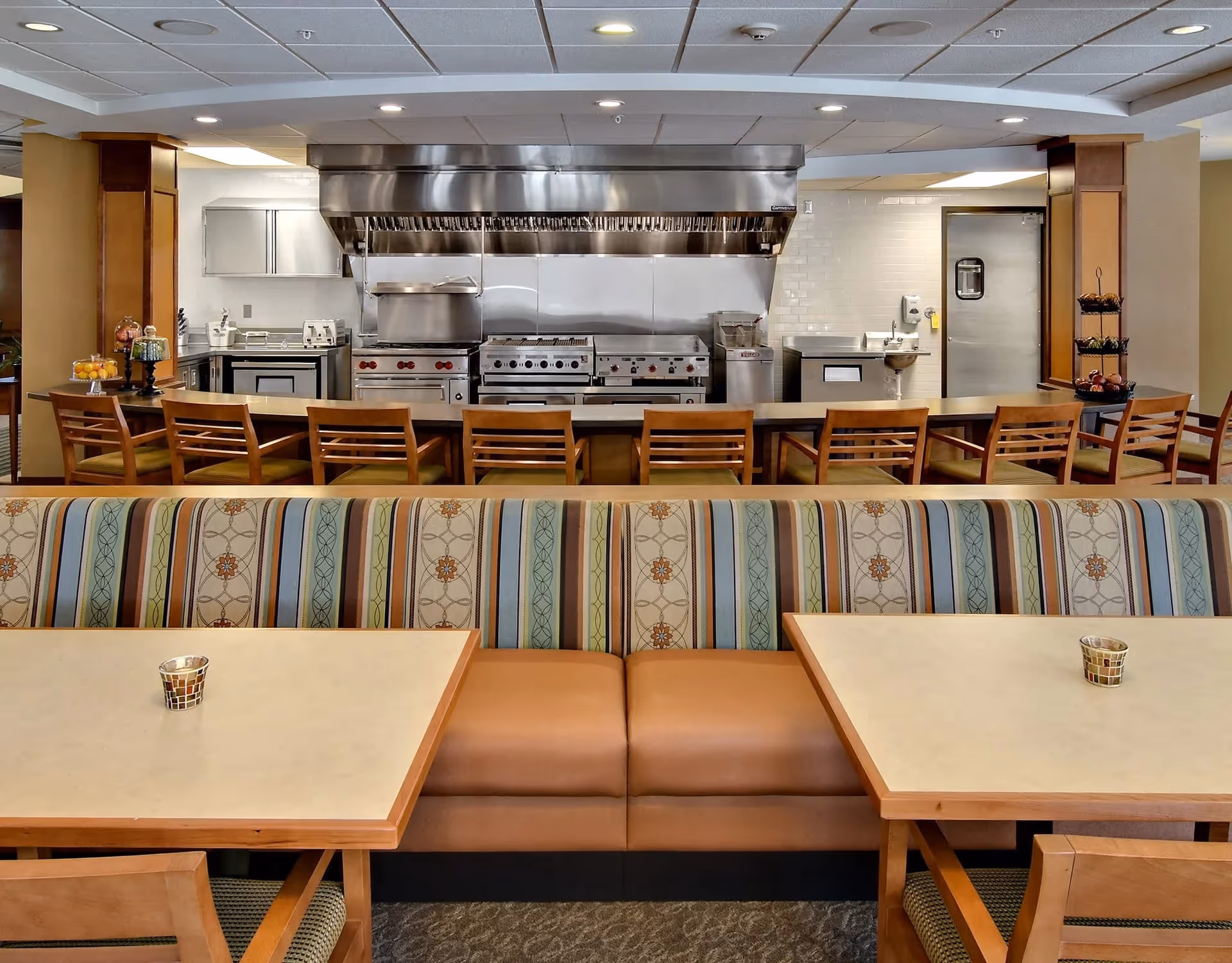 Interior view of a dining area in a senior living facility featuring a long counter with wooden chairs facing a commercial kitchen with stainless steel appliances and a large ventilation hood. In the foreground, there are cushioned booth seats with patterned upholstery and two tables, each with a small decorative candle holder.