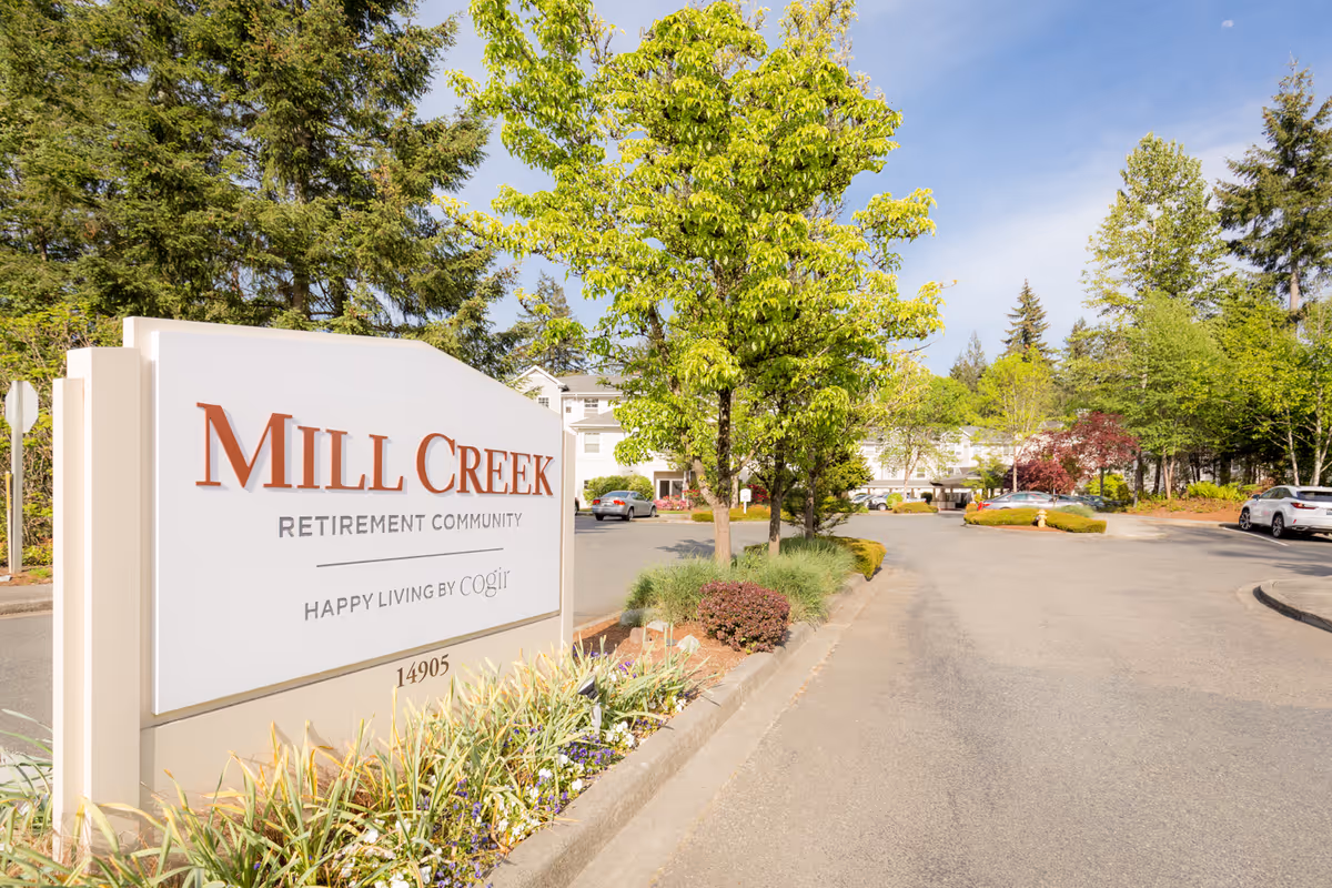 Entrance sign for Mill Creek Retirement Community with trees, shrubs, and a driveway leading to the building in the background under a clear sky.