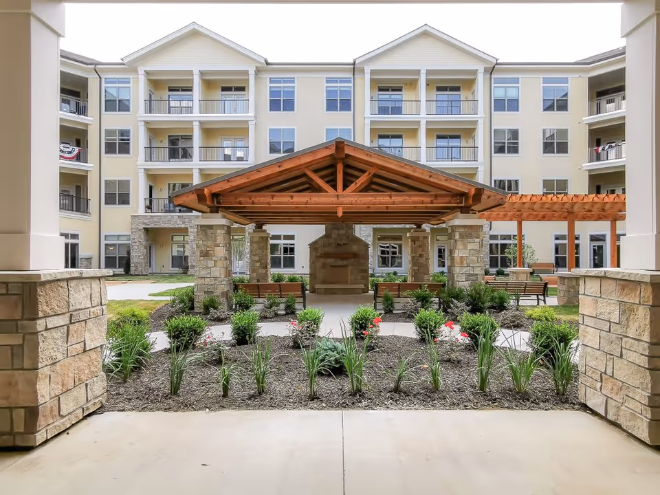 View of an outdoor courtyard area at Tiffany Springs Senior Living featuring a wooden pavilion with stone pillars, benches, landscaped garden beds with green plants and flowers, and a multi-story residential building in the background.