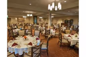 Large formal dining room with multiple round tables set with white tablecloths, folded napkins, and chairs under chandeliers.