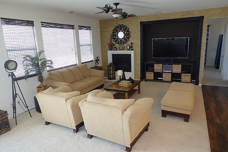 A cozy living room with beige sofas and an ottoman arranged around a wooden coffee table. The room features a fireplace with decorative items on the mantel, a wall-mounted flat-screen TV above a storage unit with wicker baskets, three windows with bamboo blinds, a standing clock, and a potted plant. The floor is carpeted with a section of wood flooring visible near the hallway.