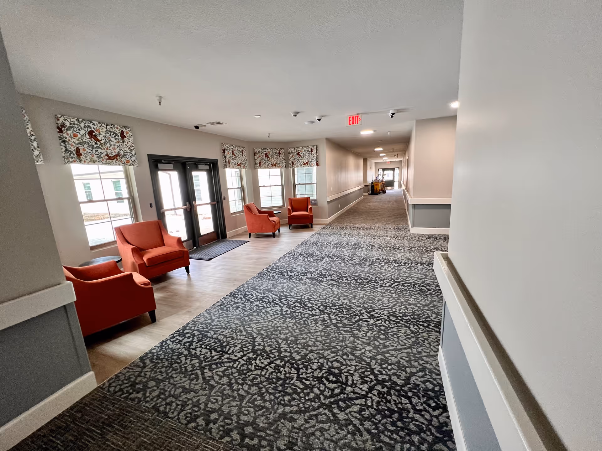 A long, carpeted hallway in a senior living facility with light gray walls and white trim. On the left side near the entrance, there are four red armchairs arranged near large windows with floral patterned valances. Double glass doors lead outside. The hallway extends into the distance with an exit sign visible on the ceiling.